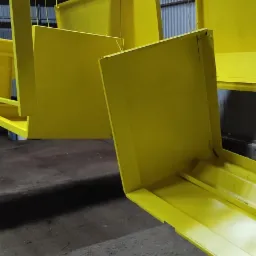 Żółte, metalowe elementy zawieszone na hakach pod czerwonym, metalowym stelażem, prawdopodobnie po procesie lakierowania.