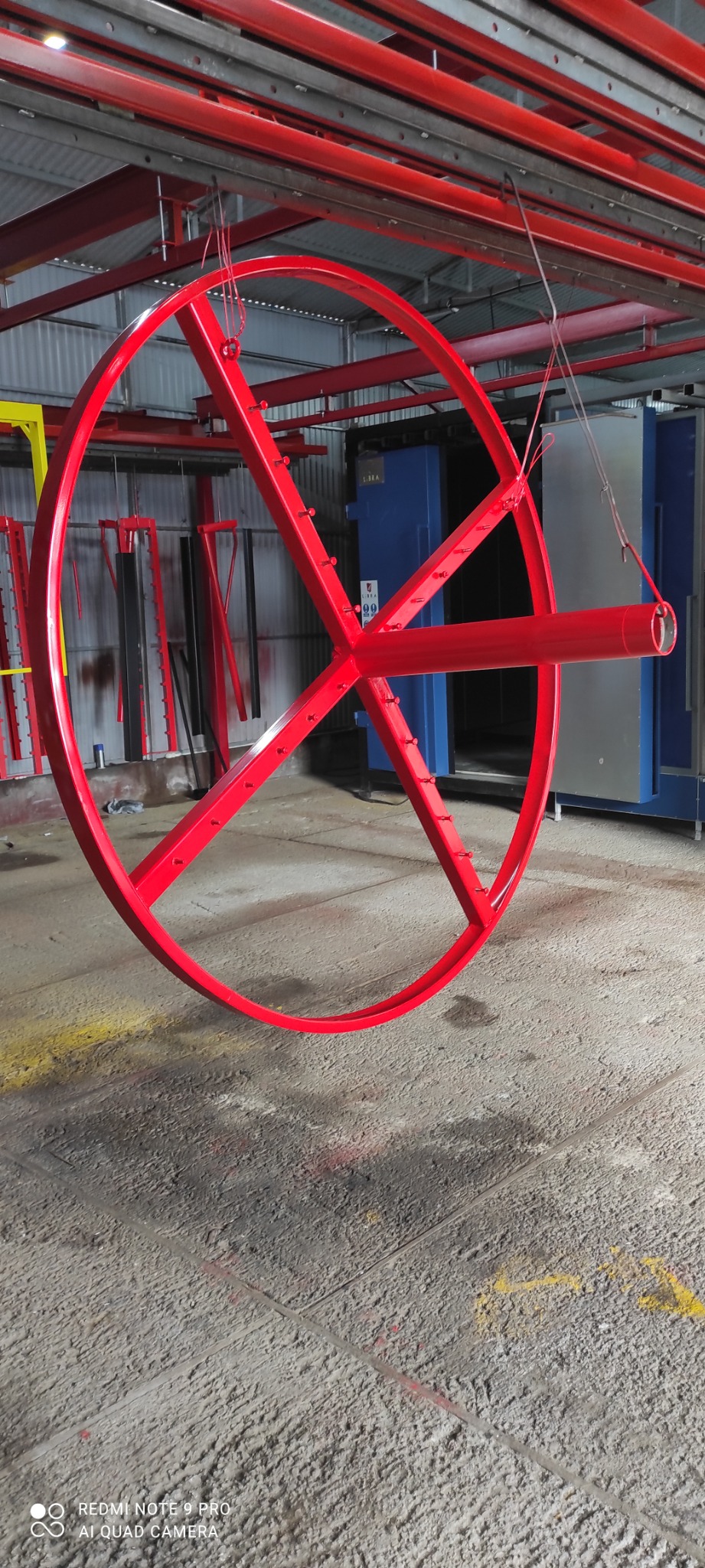Czerwone, metalowe koło z poprzeczkami zawieszone w hali produkcyjnej, prawdopodobnie po procesie malowania; widoczne ślady farby na podłodze.