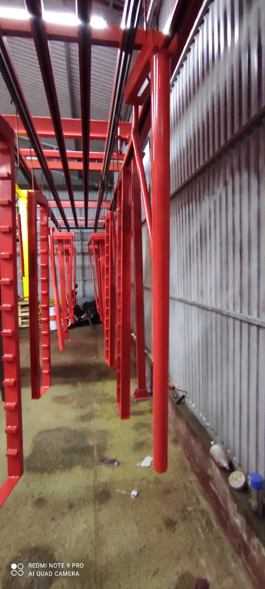 Czerwone metalowe konstrukcje, prawdopodobnie ramy, wiszące na szynach w hali w Gdyni, po procesie malowania. Świeża, intensywna, błyszcząca czerwona farba pokrywa elementy.