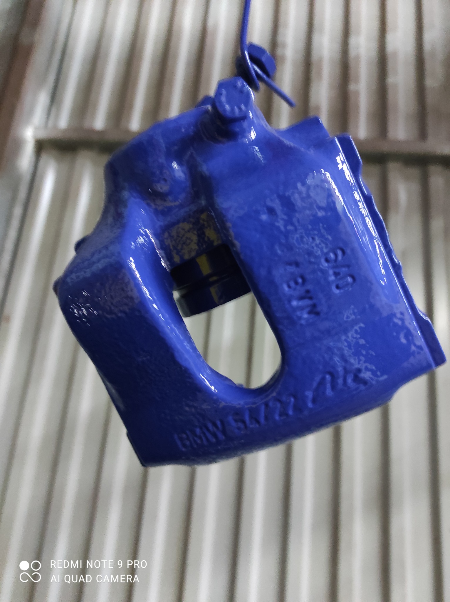 Niebieski zacisk hamulcowy BMW, świeżo po nałożeniu powłoki, zawieszony na drutu, z widocznymi oznaczeniami modelu na powierzchni.