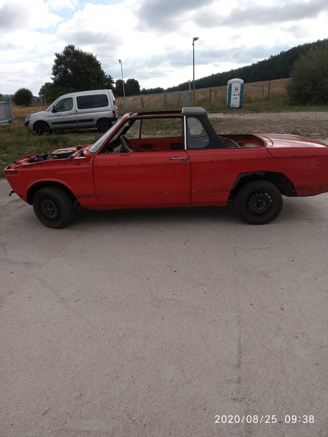 Czerwony, zdekompletowany samochód bez maski i z widocznymi śladami korozji, stojący na betonowym podłożu, z białym vanem i przenośną toaletą w tle.