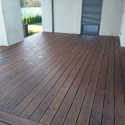 Nowo wykonany taras z desek kompozytowych w odcieniach brązu, z widoczną strukturą drewna, na tle białego budynku z szarą roletą zewnętrzną i widokiem na zielony ogród.