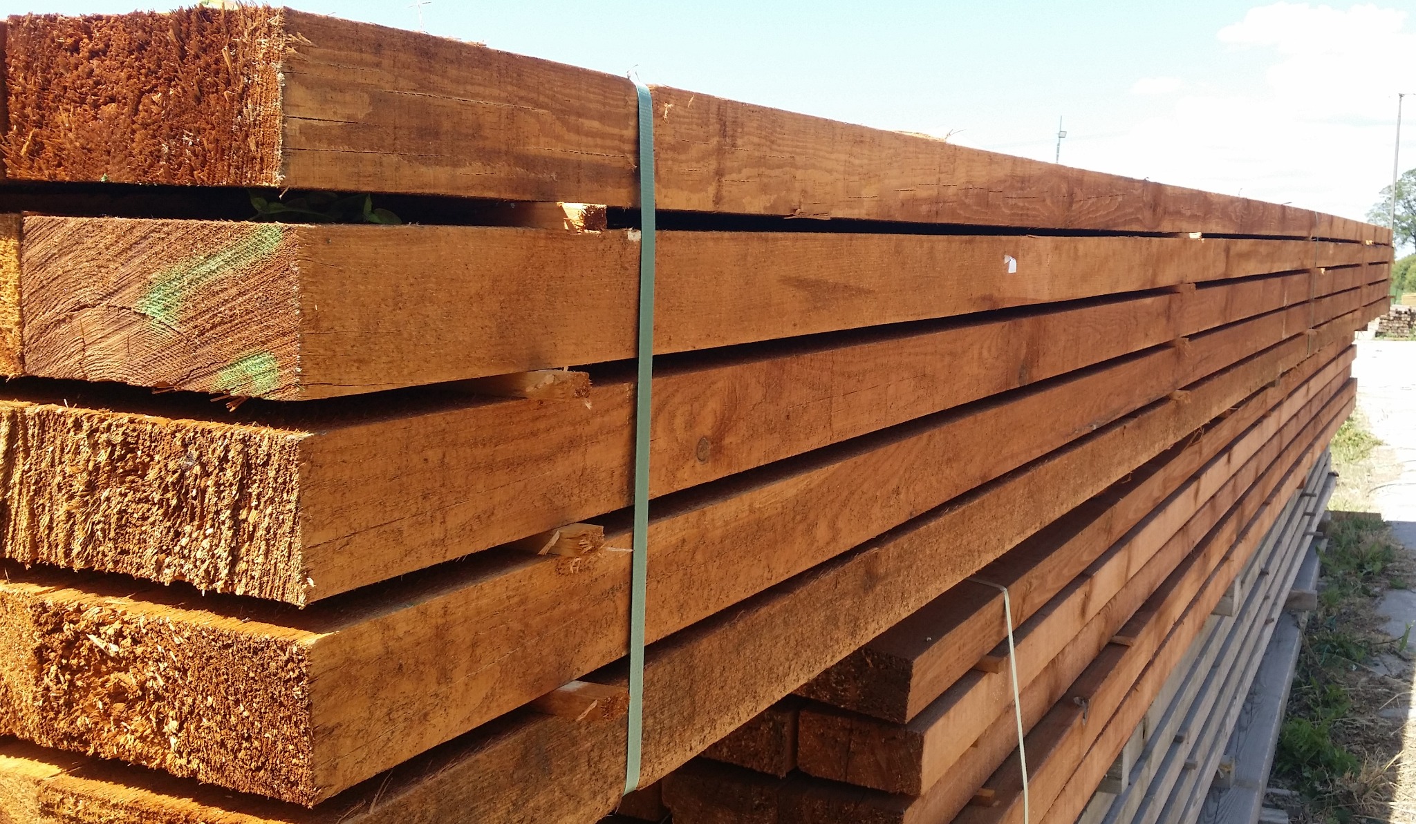 Stos brązowych, prostokątnych belek drewnianych spiętych zieloną taśmą, widoczne ślady po cięciu i zielone oznaczenia na niektórych elementach, ułożonych na paletach na zewnątrz.