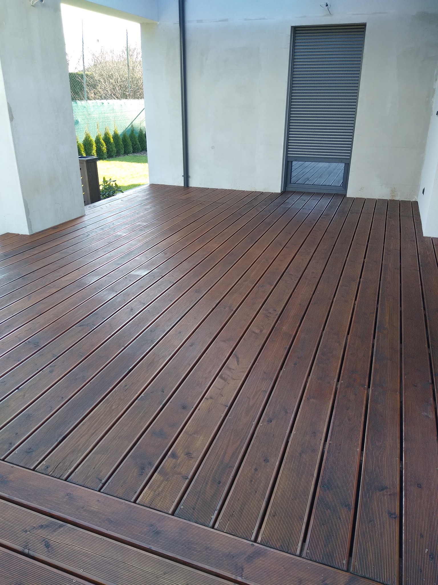 Nowo wykonany taras z desek kompozytowych w odcieniach brązu, z widoczną strukturą drewna, na tle białego budynku z szarą roletą zewnętrzną i widokiem na zielony ogród.