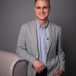 Uśmiechnięty mężczyzna w szarej marynarce w jodełkę i błękitnej koszuli, opierający się o beżowy fotel na szarym tle.