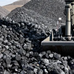 Kopuła czarnego węgla kamiennego i jasnoszarych hałd w tle, z mechaniczną łopatą koparki zanurzoną w węglu.