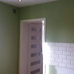 Świeżo pomalowane na zielono wnętrze z białą, częściowo wykończoną ścianą z płytek imitujących cegłę, białe drzwi z przeszkleniami i drewniana podłoga.