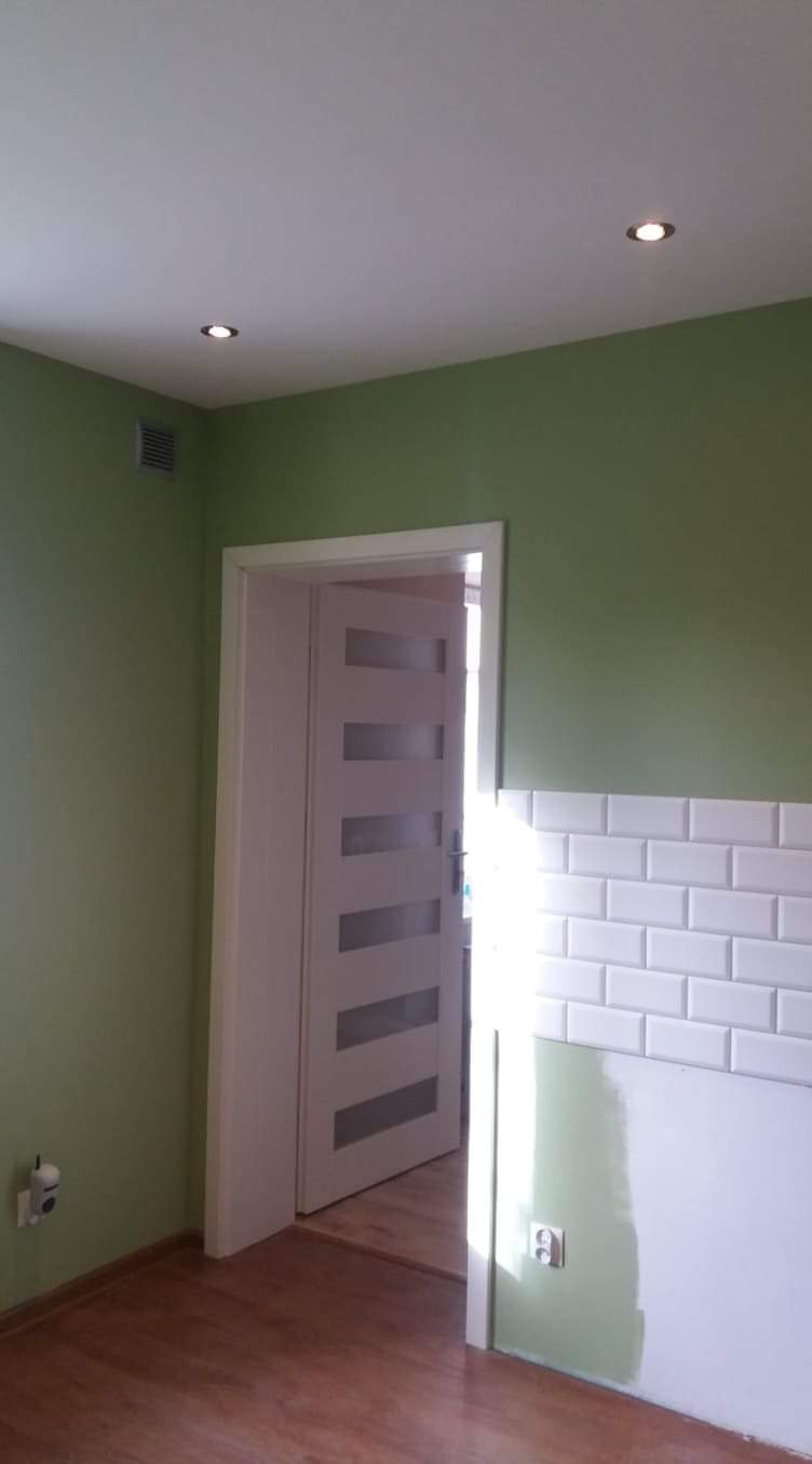 Świeżo pomalowane na zielono wnętrze z białą, częściowo wykończoną ścianą z płytek imitujących cegłę, białe drzwi z przeszkleniami i drewniana podłoga.