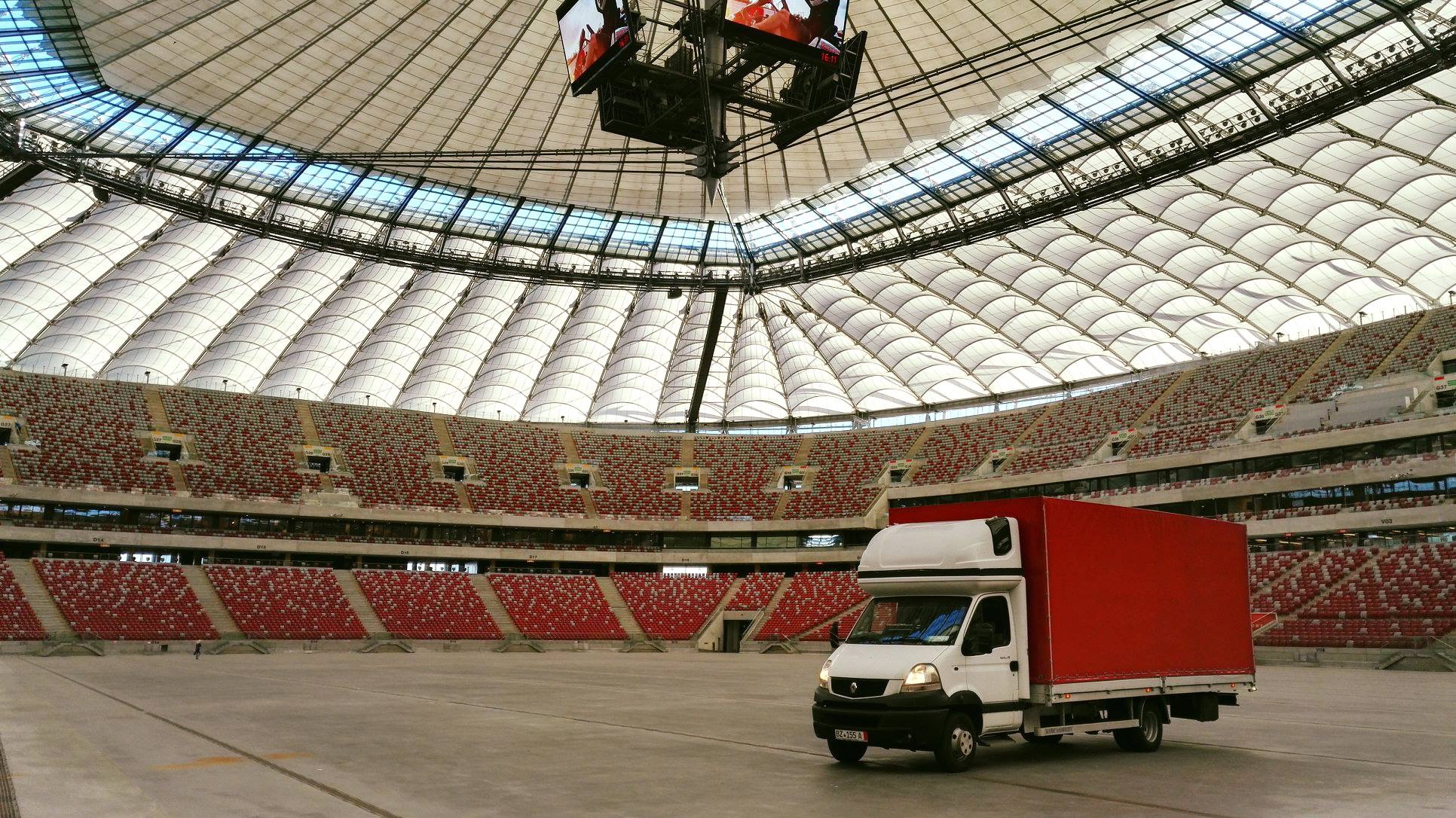 Czerwony samochód dostawczy na płycie stadionu Narodowego w Warszawie, widok na puste trybuny i konstrukcję dachu.