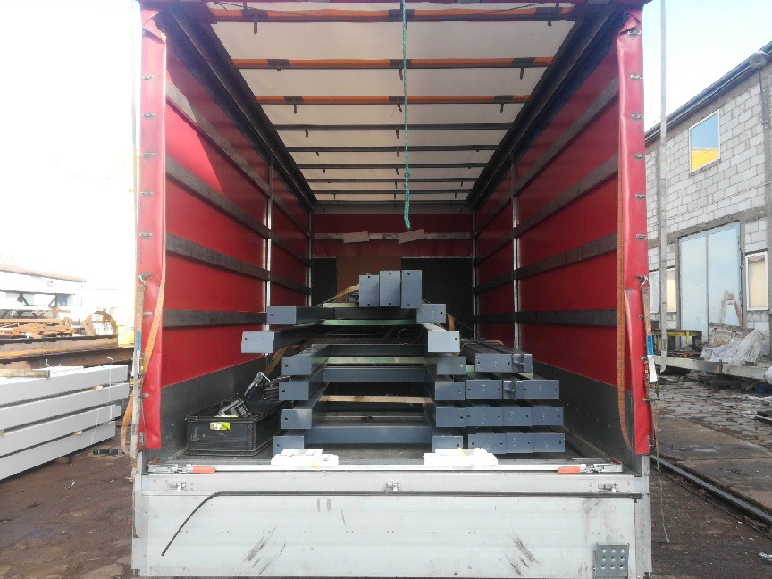 Wnętrze naczepy ciężarówki z czerwonymi plandekami bocznymi, załadowanej szarymi, stalowymi elementami konstrukcyjnymi, widok z tyłu pojazdu, w tle budynek z oknem z ukraińską flagą.