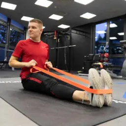 Mężczyzna w czerwonej koszulce ćwiczy rozciąganie z gumą oporową siedząc na macie w siłowni w Warszawie. W tle widoczne sprzęty do ćwiczeń.