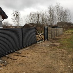 NAGET Nowoczesne Systemy Ogrodzeń Monika Naguszewska - Nowoczesna, przesuwna brama wjazdowa z czarnych paneli, częściowo zamontowana, z widocznymi narzędziami (młotek, klucz) na błotnistym terenie działki, w tle dom i drzewa pod pochmurnym niebem.