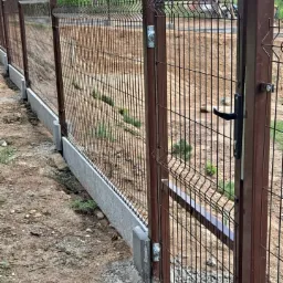 Brązowa brama wjazdowa z panelem siatkowym, widoczne betonowe podmurówki i słupki, teren po instalacji w trakcie wyrównywania.