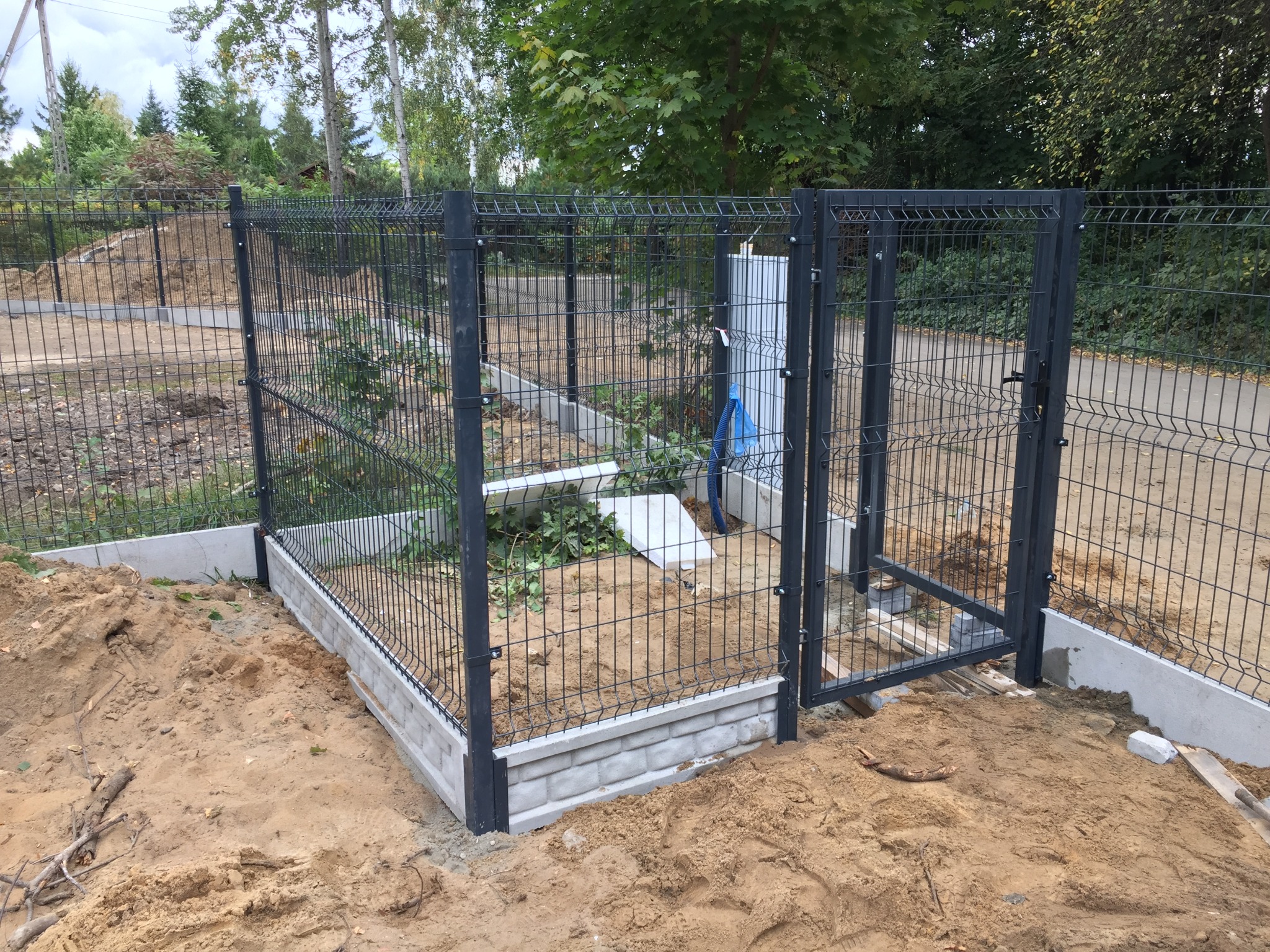 Ciemnoszare ogrodzenie panelowe z bramą wjazdową, osadzone na betonowej podmurówce, teren wokół w trakcie prac ziemnych, widoczne ślady piasku i wykopów.