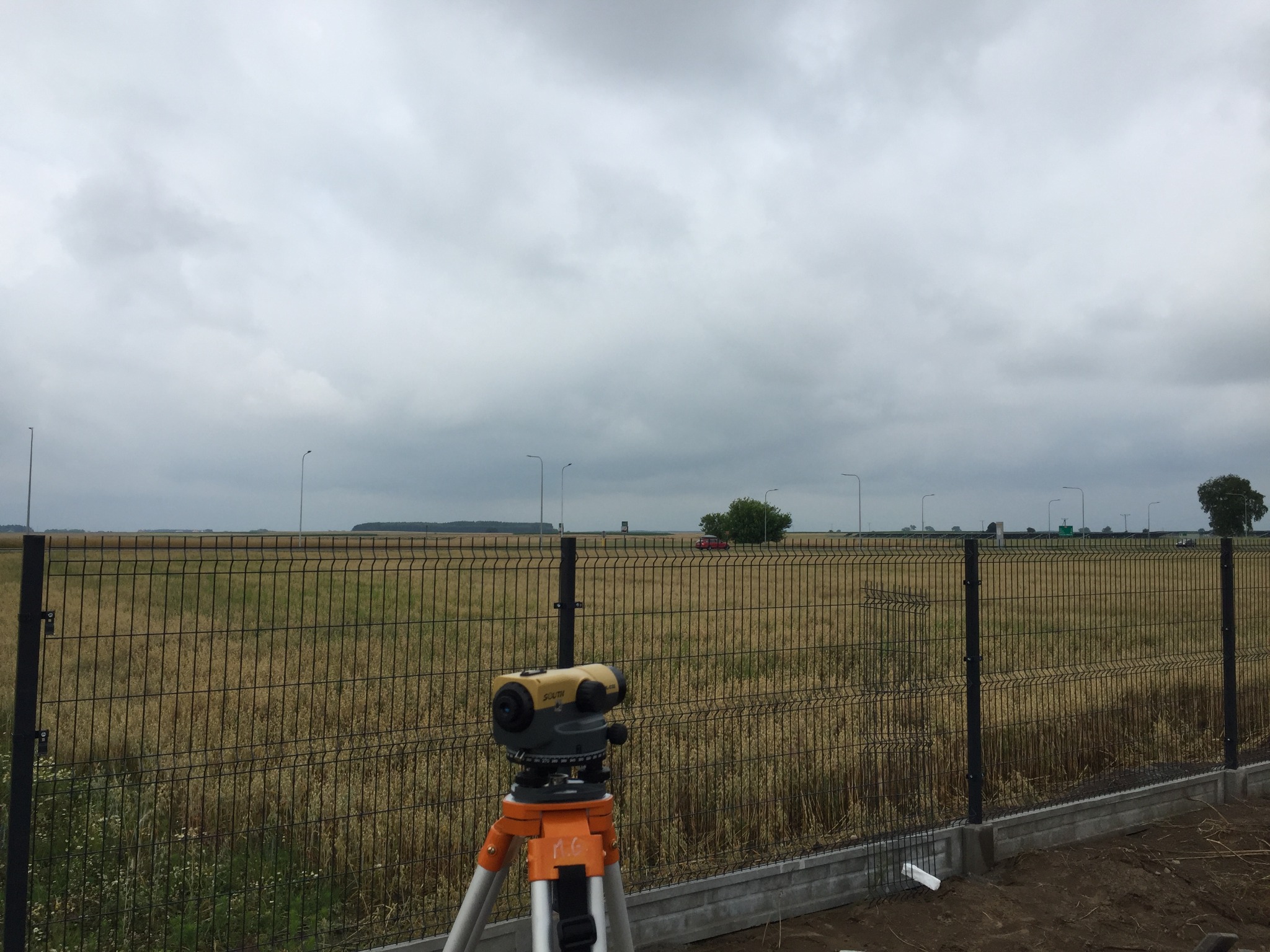 Pomiar geodezyjny nowo postawionego ogrodzenia panelowego w Ciechanowie, widoczny niwelator na statywie, w tle droga i pochmurne niebo.