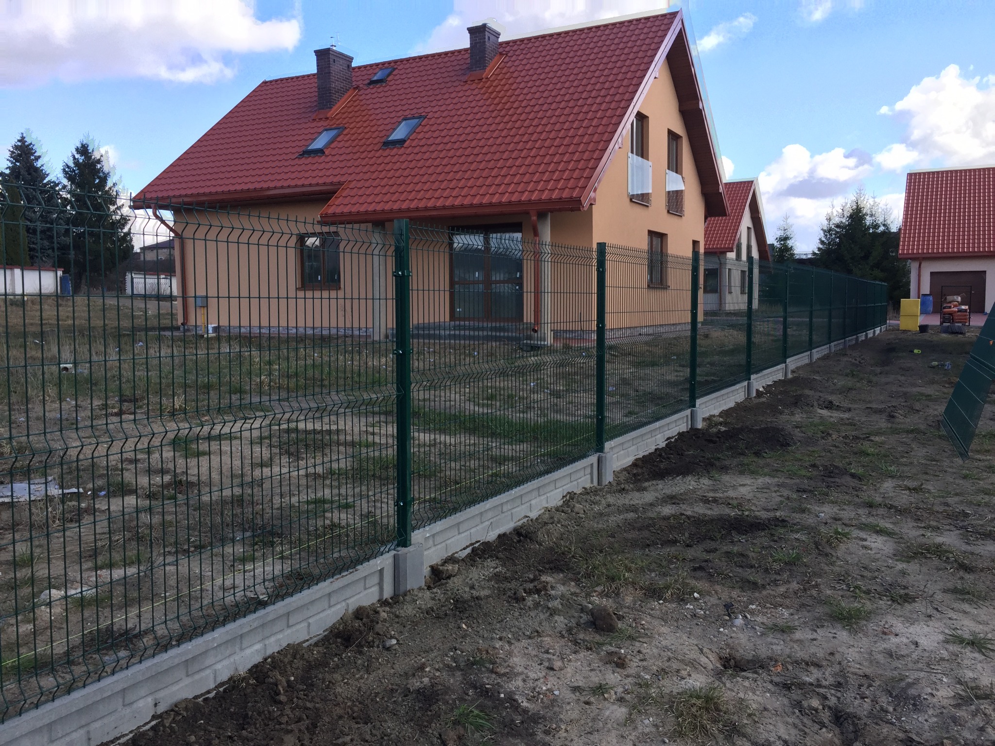 Zielone ogrodzenie panelowe na betonowej podmurówce otacza szereg domów z czerwoną dachówką w słoneczny dzień.