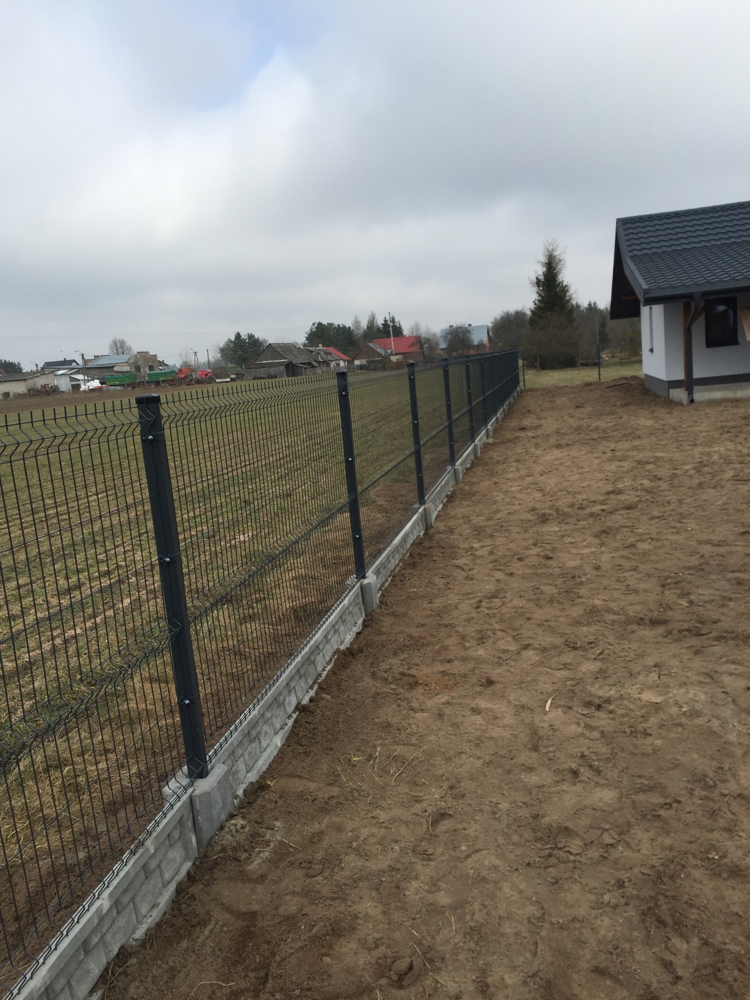 Nowoczesne czarne ogrodzenie panelowe na betonowej podmurówce, widok na dom z szarą dachówką i pole w tle, pochmurne niebo.