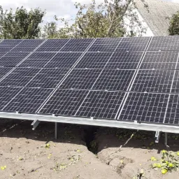 Instalacja paneli słonecznych na metalowej konstrukcji w ogrodzie, widoczne jabłka pod panelami i dom w tle.