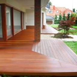 Kostroma - Drewniany taras z desek ułożonych wzdłuż, z niskimi schodami i kolumną z cegły klinkierowej, widok na ogród z drzewami i dom z czerwoną dachówką.