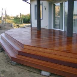 Kostroma - Nowoczesny taras z drewna egzotycznego z trzema stopniami, lśniący po impregnacji, z widocznymi betonowymi podstawami i fragmentem domu z szarymi kolumnami.