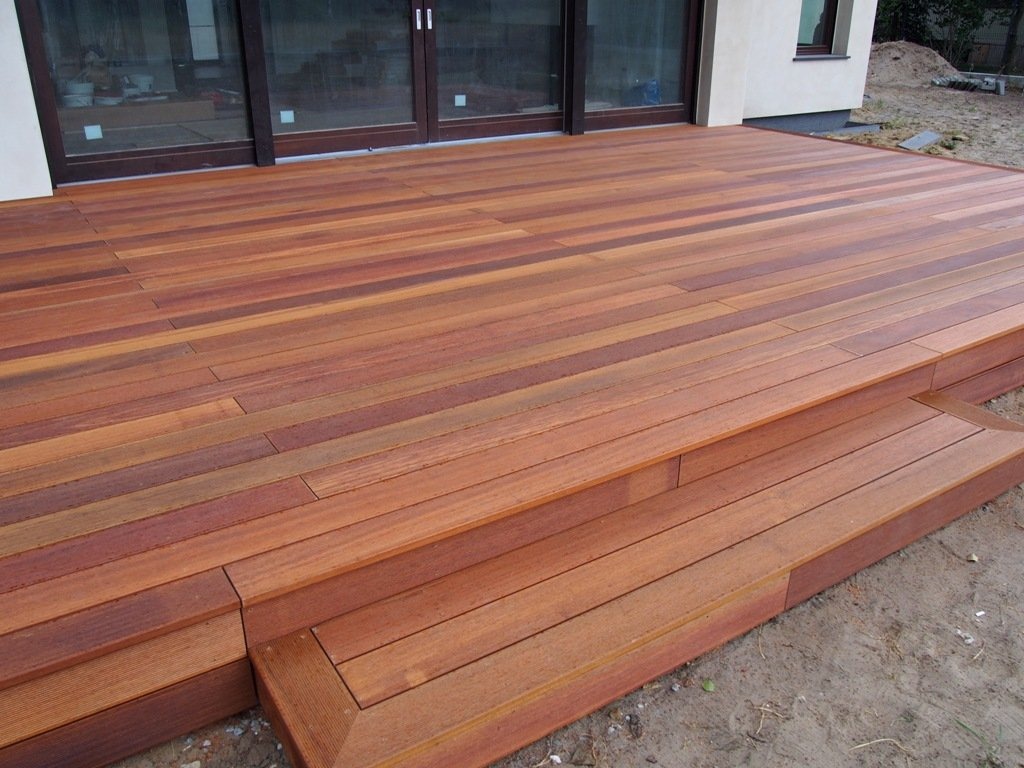 Nowoczesny taras z desek o ciepłej, czerwono-brązowej barwie, prowadzący do przeszklonych drzwi balkonowych. Widoczne schodki tarasowe z wyraźnym obramowaniem. W tle fragment elewacji budynku.