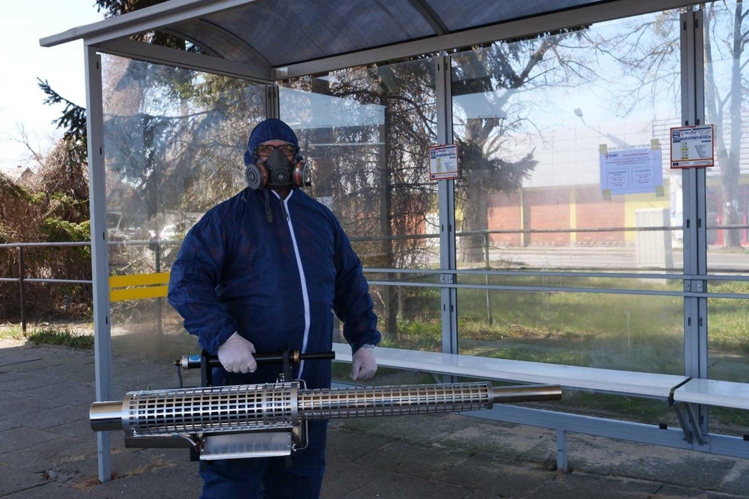 Osoba w kombinezonie ochronnym i masce przeciwgazowej, trzymająca metalowy opryskiwacz w przystanku autobusowym.
