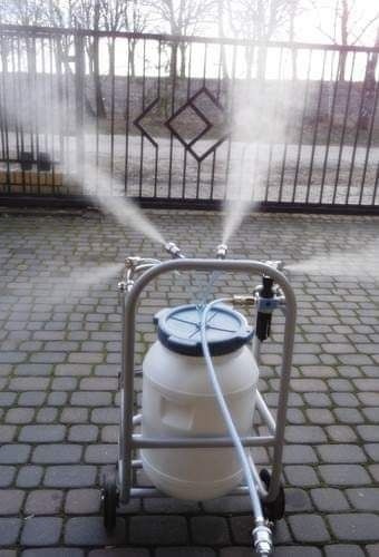 Mobilne urządzenie do zamgławiania z białym zbiornikiem i metalową ramą na kółkach, rozpylające wodę przed metalową bramą z ozdobnym wzorem.