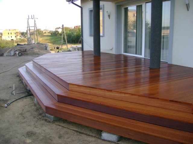 Nowoczesny taras z drewna egzotycznego z trzema stopniami, lśniący po impregnacji, z widocznymi betonowymi podstawami i fragmentem domu z szarymi kolumnami.