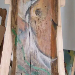 Portret kobiety namalowany na kawałku tarcicy, wsparty na drewnianym stojaku, z zielonymi akcentami roślinnymi.