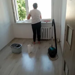 Kobieta w trakcie mycia okna w jasnym pokoju z panelami, widoczny mop, wiadro i odkurzacz.