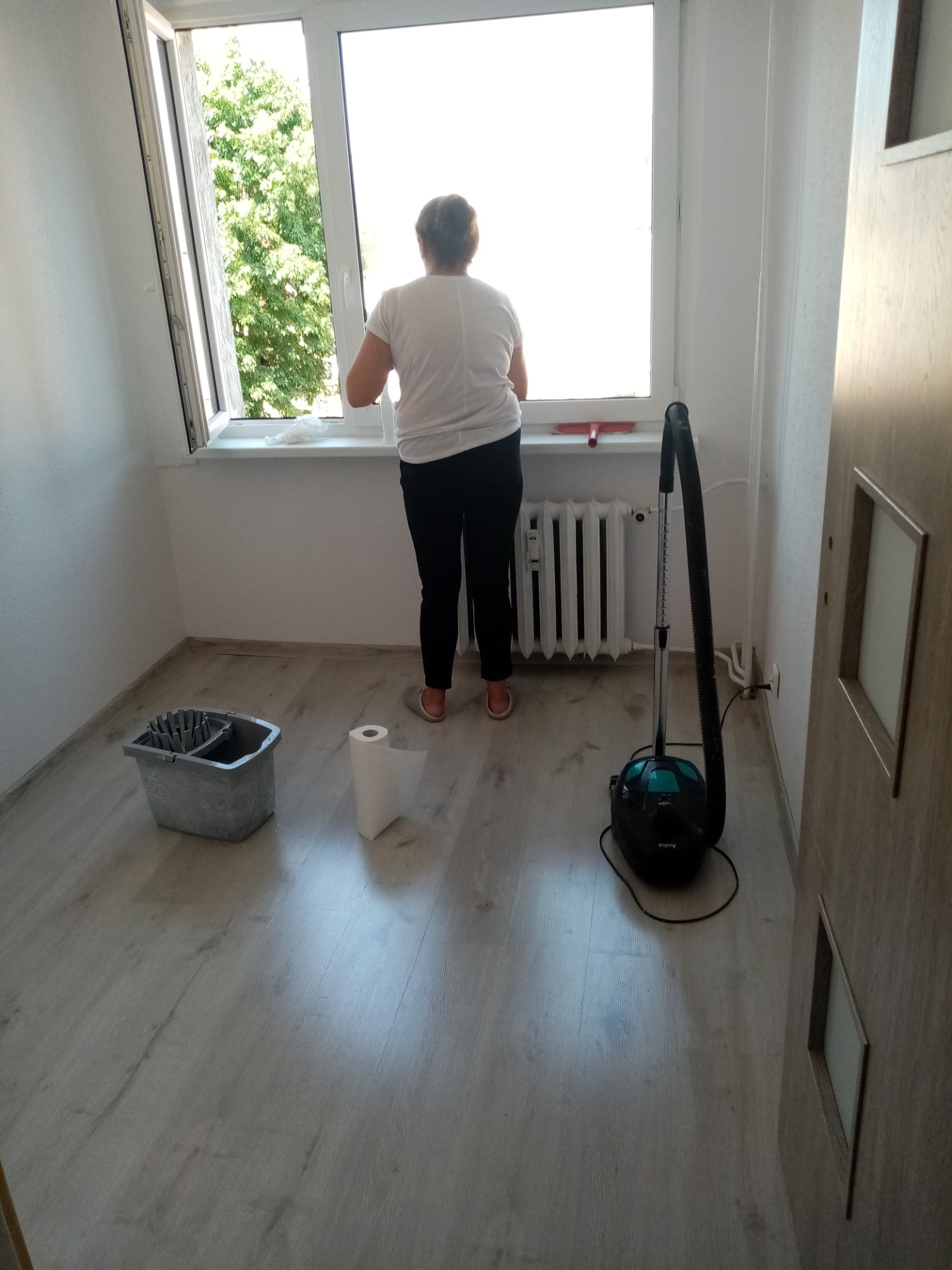 Kobieta w trakcie mycia okna w jasnym pokoju z panelami, widoczny mop, wiadro i odkurzacz.