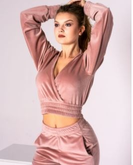 Kobieta w welurowym komplecie dresowym w kolorze pudrowego różu, bluzka z dekoltem w serek i długimi rękawami, spodnie z kieszeniami, włosy upięte w wysoki kucyk, neutralne tło.