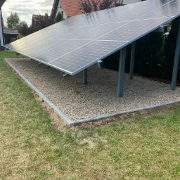 Panel słoneczny na metalowej konstrukcji, umieszczony na żwirowym podłożu z obrzeżem, trawnik w tle, widok z poziomu gruntu.