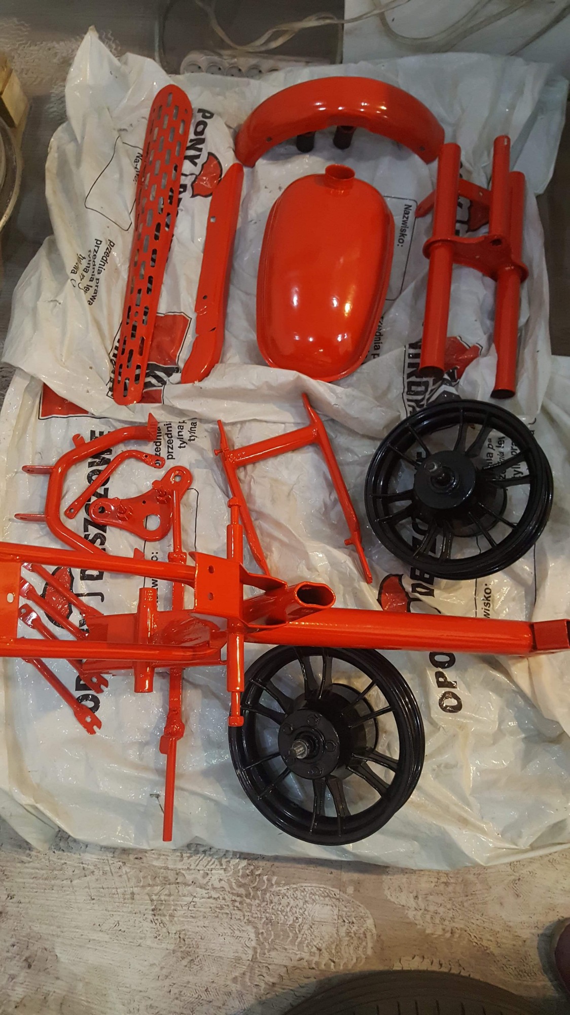 Części motocykla po renowacji i pomalowaniu na jaskrawy czerwony kolor, leżące na białym worku foliowym. Widoczne elementy to błotnik, zbiornik paliwa, rama, siedzenie i koła.