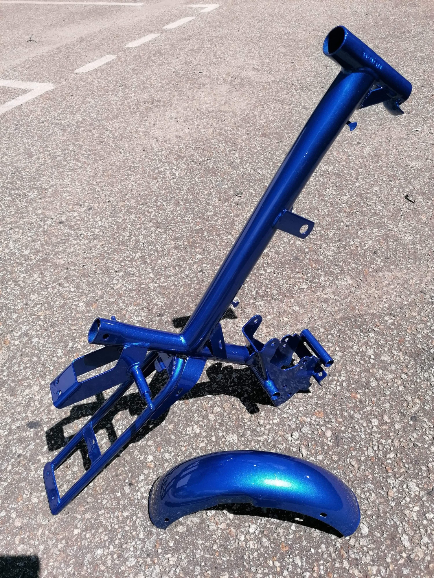 Rama i błotnik motocyklowy po renowacji, pokryte błyszczącą, niebieską farbą proszkową, leżące na szorstkim, asfaltowym podłożu w jasnym świetle słonecznym.