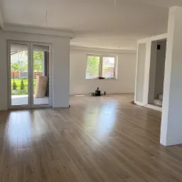 Jasne, przestronne wnętrze w trakcie prac wykończeniowych z drewnopodobną podłogą, świeżo pomalowanymi ścianami i widocznymi elementami instalacji elektrycznej.