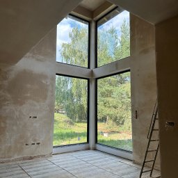 Kuba - Wnętrze domu w trakcie tynkowania maszynowego, widoczne duże okno z widokiem na zieleń, ściany pokryte surowym tynkiem, podłoga zabezpieczona folią, drabina oparta o ścianę.