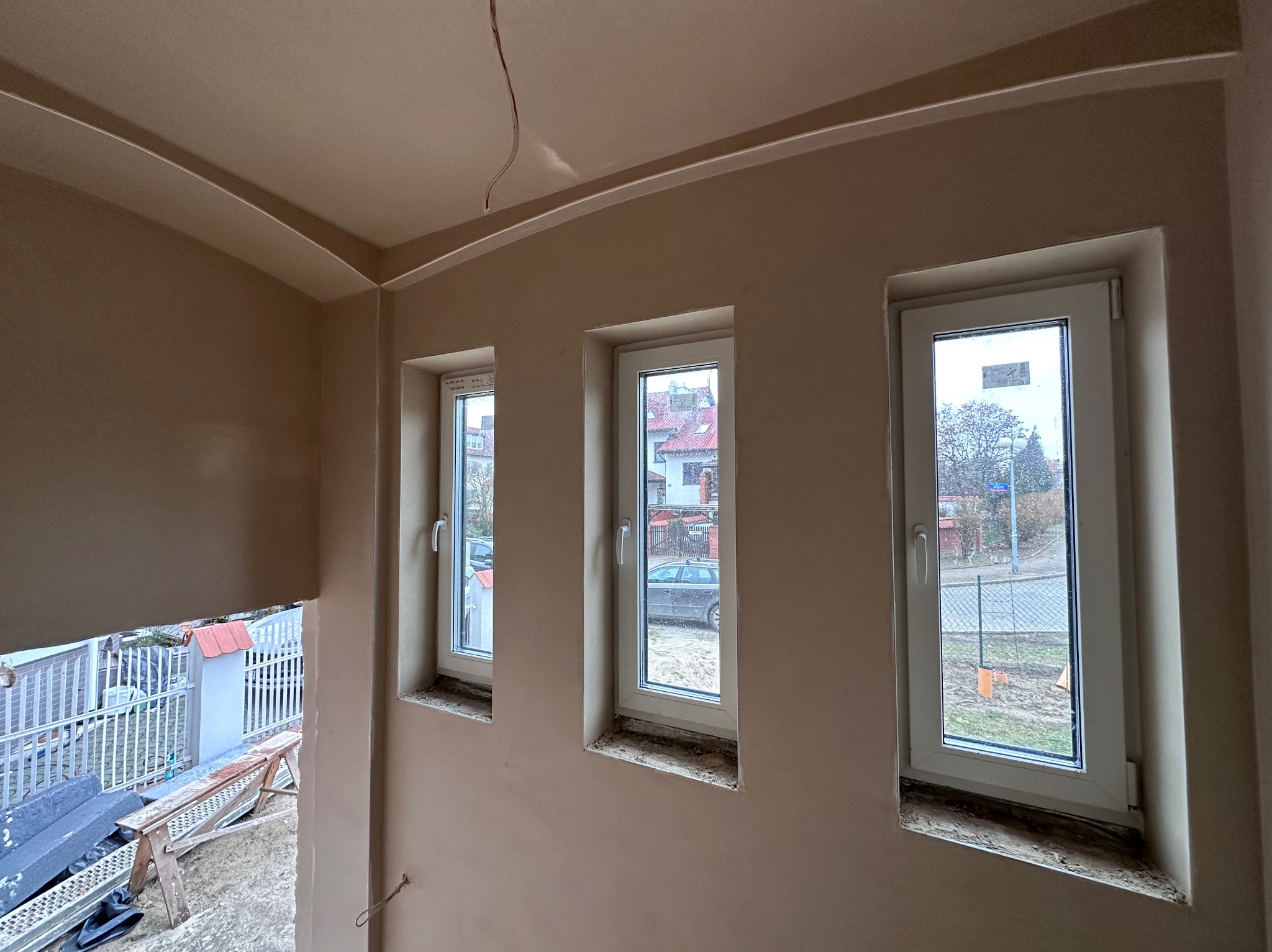 Wnętrze po nałożeniu jasnego tynku maszynowego, widoczne trzy okna z białymi ramami i zaokrąglone wykończenie sufitu. Na zewnątrz widok na ulicę i domy.