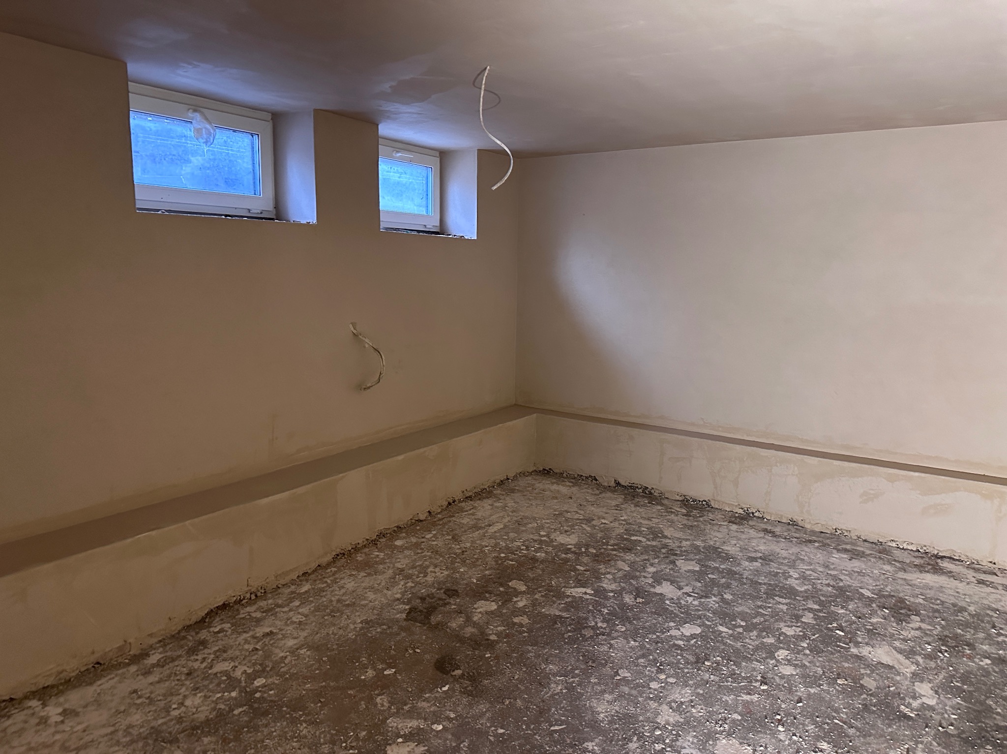 Pokój po nałożeniu gładzi gipsowej na ściany, widoczne dwa małe okna i wystające przewody ze ściany i sufitu, podłoga w trakcie przygotowania.