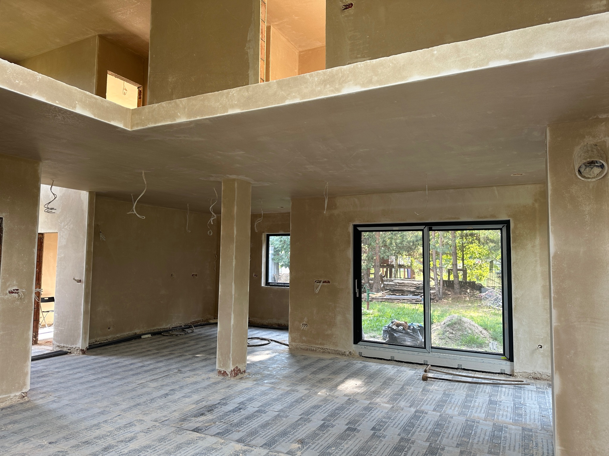 Wnętrze domu w trakcie prac wykończeniowych, ściany i sufit pokryte świeżym tynkiem, widoczne wystające przewody elektryczne, podłoga zabezpieczona folią ochronną, przez okno widok na zieleń.