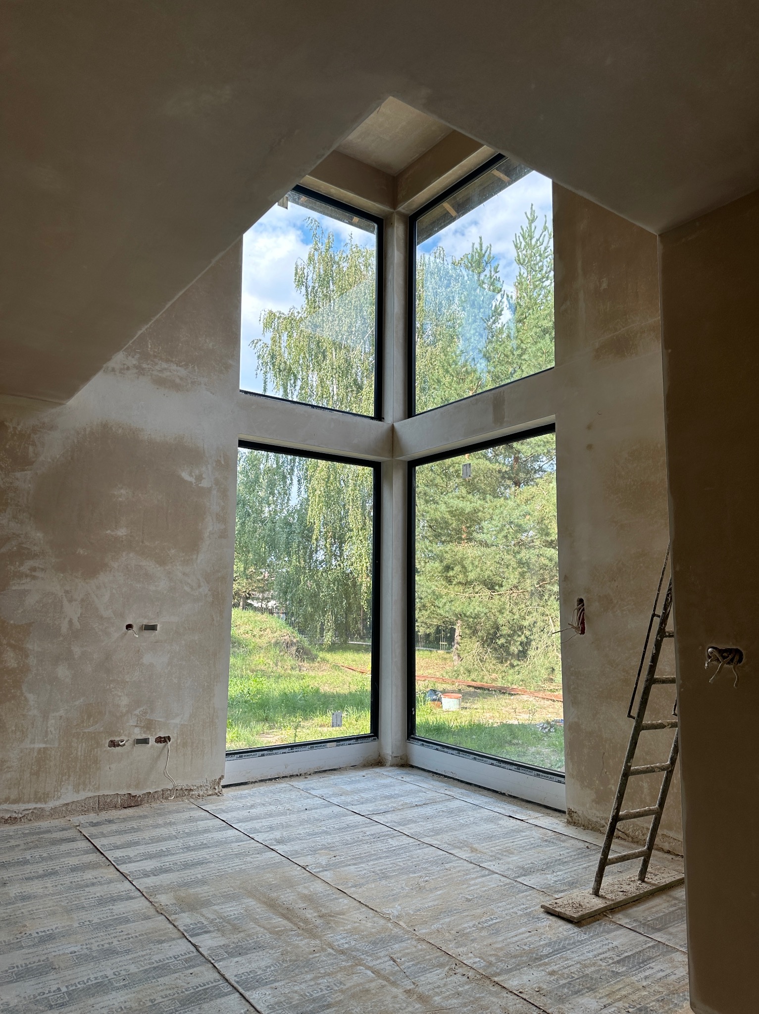 Wnętrze domu w trakcie tynkowania maszynowego, widoczne duże okno z widokiem na zieleń, ściany pokryte surowym tynkiem, podłoga zabezpieczona folią, drabina oparta o ścianę.