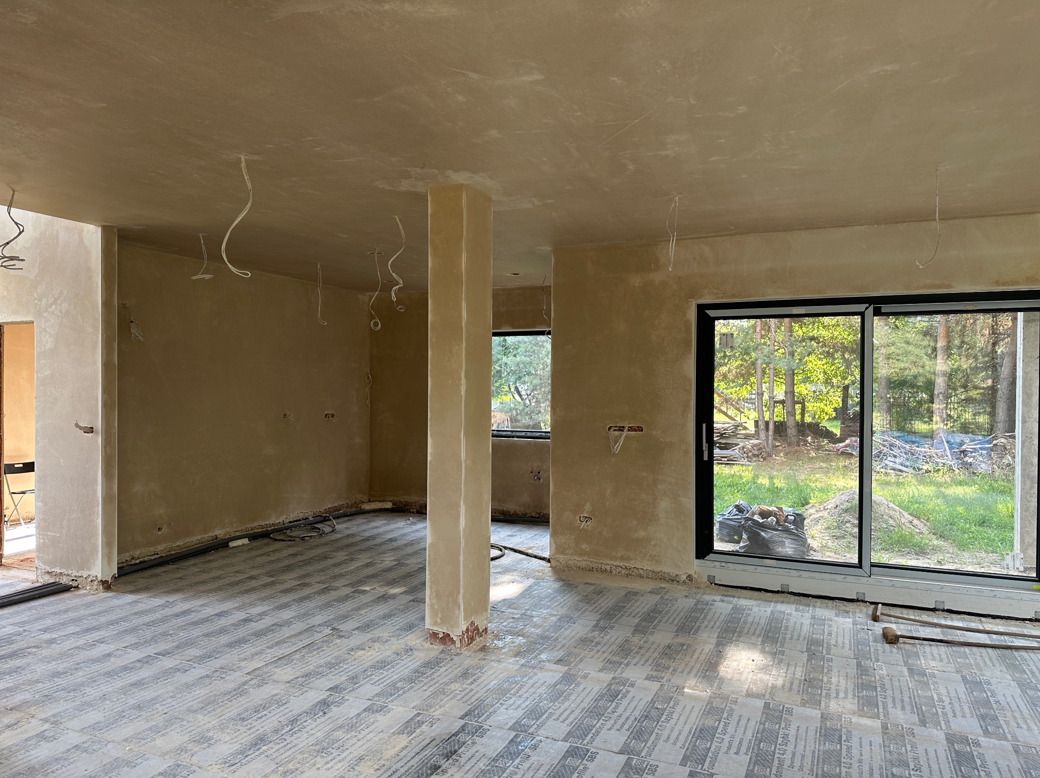 Wnętrze domu w trakcie prac wykończeniowych: ściany i sufit pokryte świeżym, jasnym tynkiem, widoczne wystające przewody elektryczne, podłoga zabezpieczona folią ochronną, duża szyba okienna...