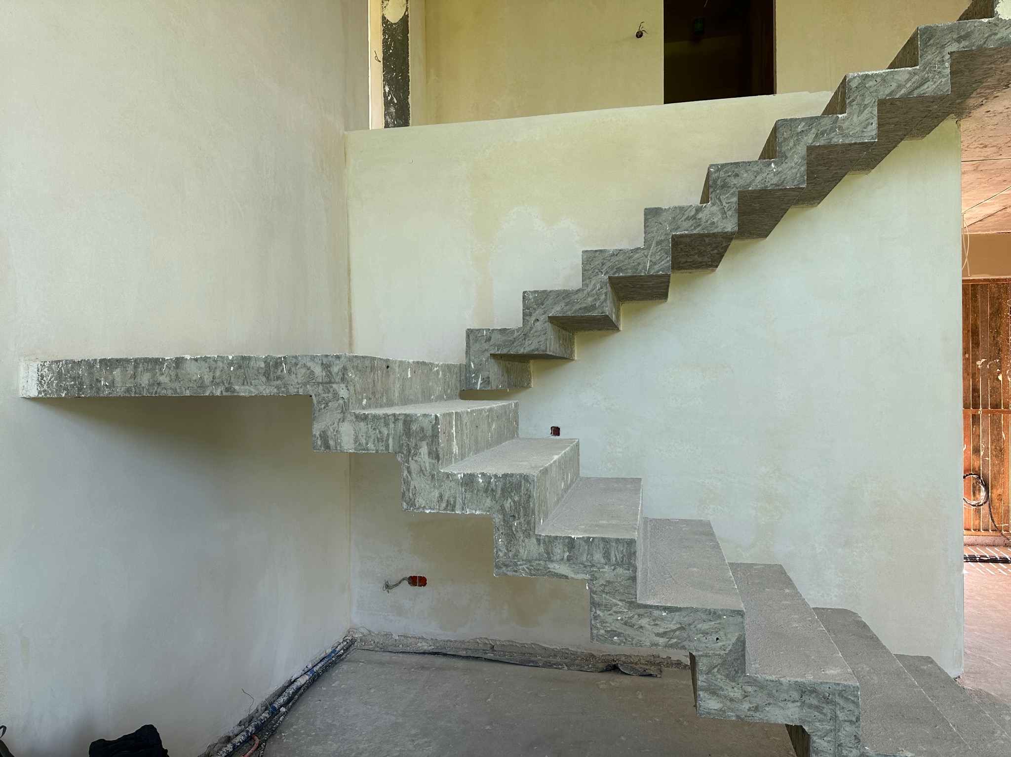 Surowe, betonowe schody wspornikowe w trakcie montażu na tle świeżo otynkowanych ścian wewnętrznych, widoczne przewody elektryczne i niedokończone wykończenie.