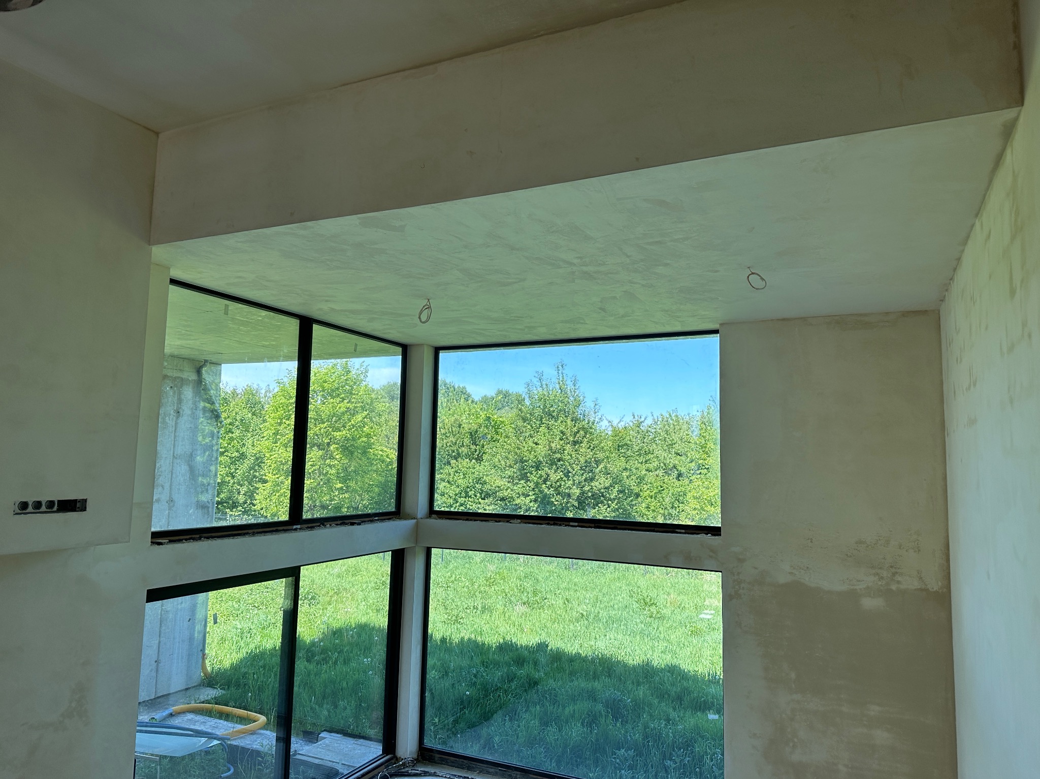 Wnętrze w stanie deweloperskim z dużymi oknami i surowymi ścianami po nałożeniu tynku, widoczne niedoskonałości i zacieki, na zewnątrz bujna zieleń.