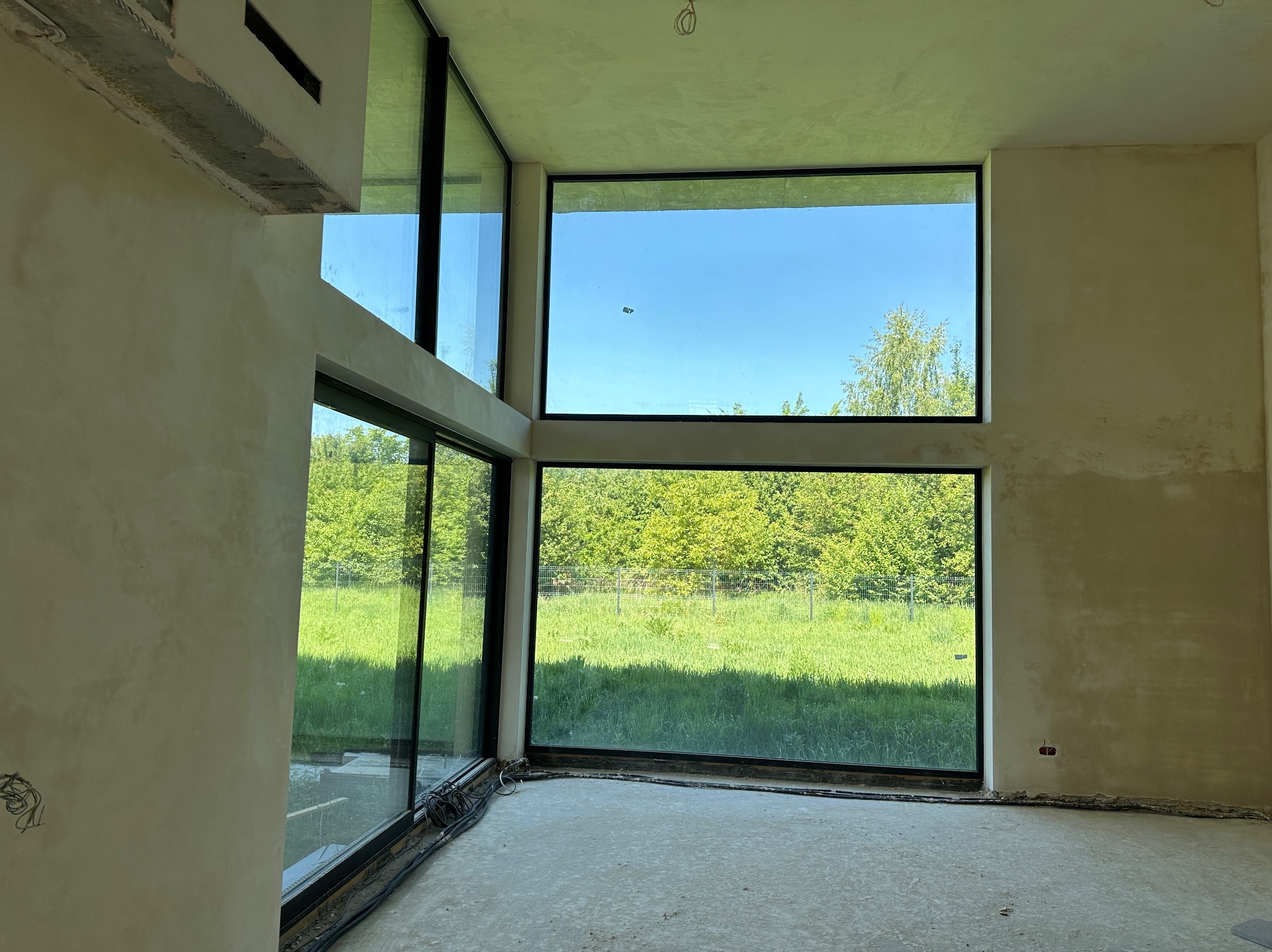 Wnętrze surowego domu z dużymi oknami, ściany świeżo pokryte tynkiem, widoczne niedoskonałości i przewody elektryczne, zieleń na zewnątrz.
