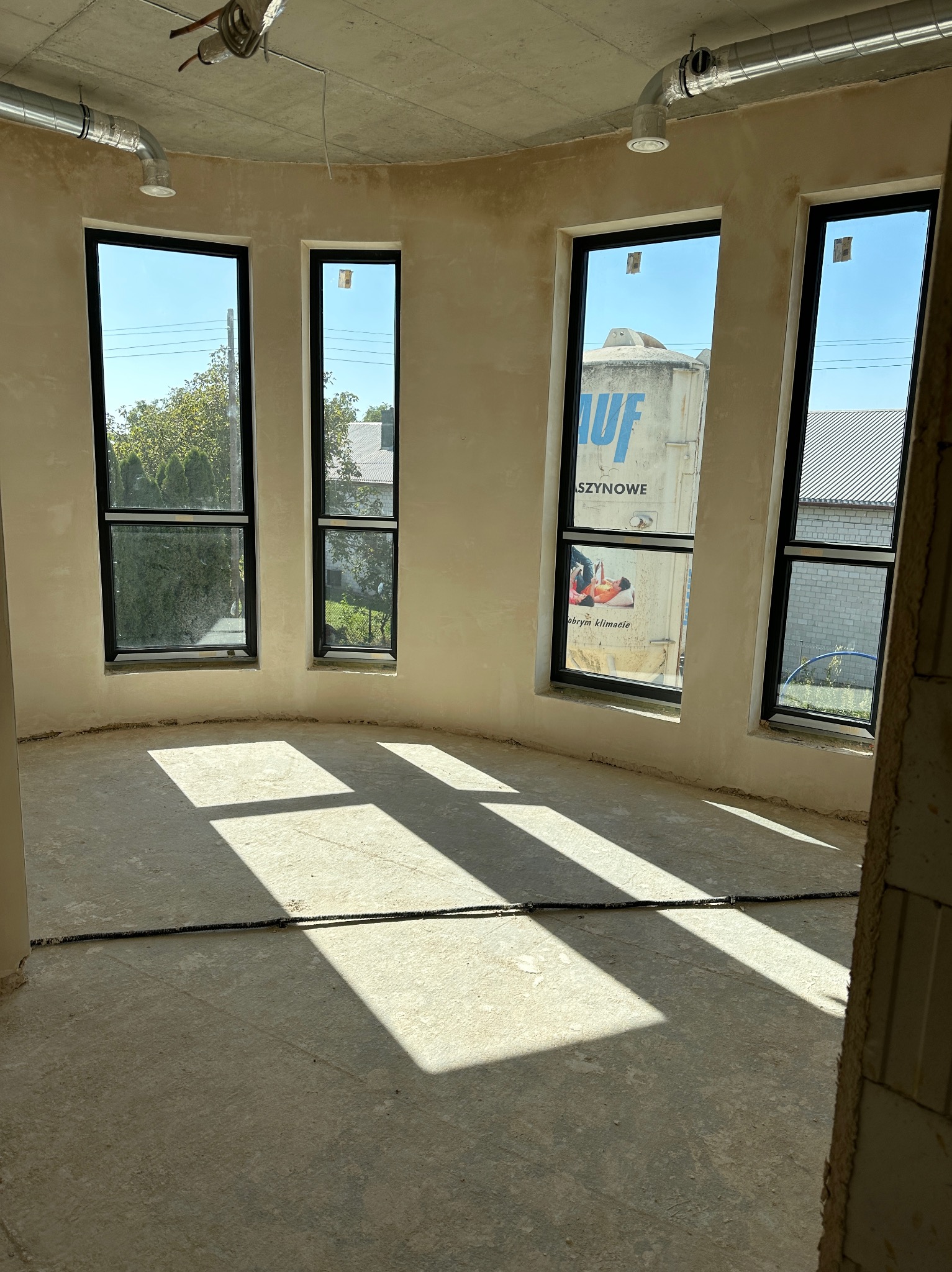 Wnętrze okrągłego pokoju w stanie surowym z trzema oknami z czarnymi ramami, na ścianach świeżo nałożone tynki, na suficie widoczne rury wentylacyjne, na podłodze długie cienie rzucane przez...