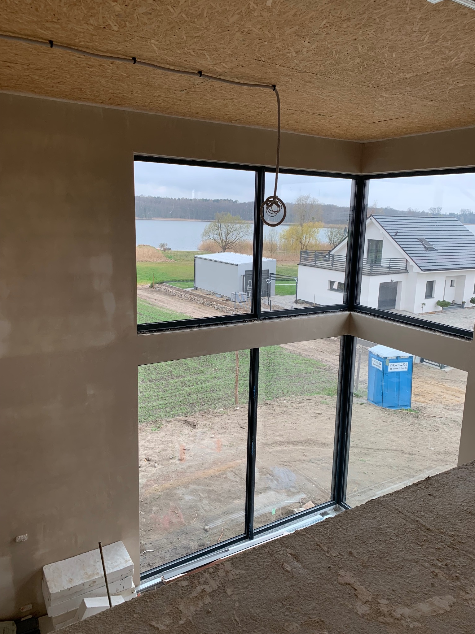 Wnętrze w stanie deweloperskim z nałożonym tynkiem na ścianach, widok na jezioro i nowo wybudowany dom przez panoramiczne okna, na zewnątrz widoczna przenośna toaleta.