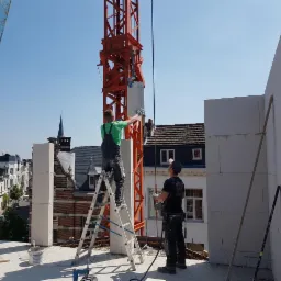 Dwóch murarzy w trakcie wznoszenia ściany z białych bloczków na dachu budynku, z użyciem dźwigu i drabiny, na tle zabudowy miejskiej z kościołem i błękitnym niebem.