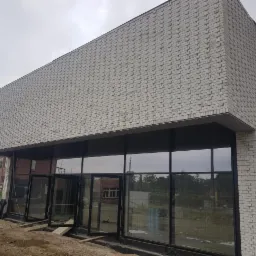 Nowoczesny budynek z białej cegły z dużymi przeszkleniami, widok z zewnątrz na placu budowy, pochmurne niebo.