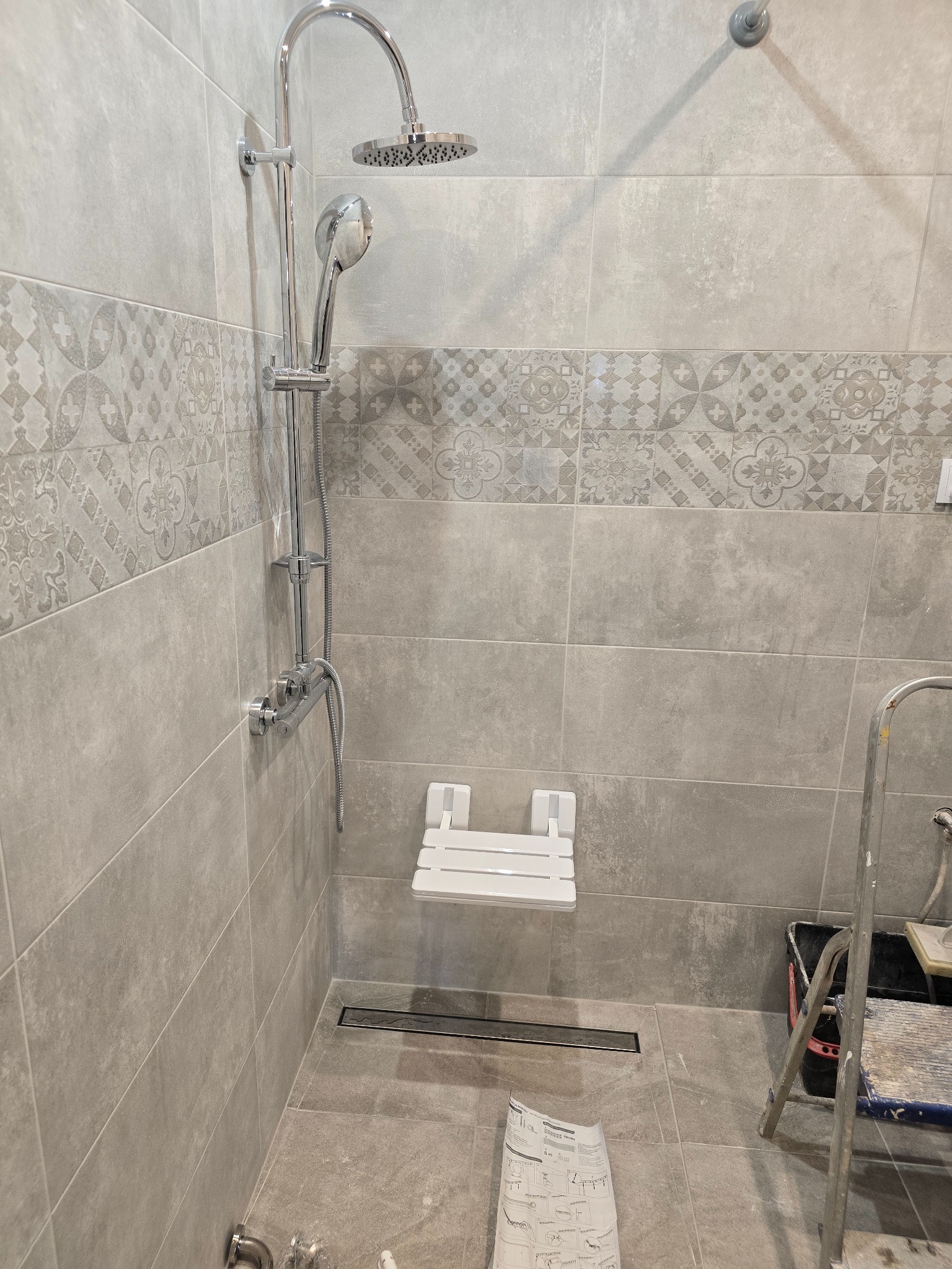 Wykończona kabina prysznicowa z szarymi płytkami, deszczownicą i siedziskiem. Widoczny odpływ liniowy i drabina malarska. Dekoracyjne płytki na ścianie.